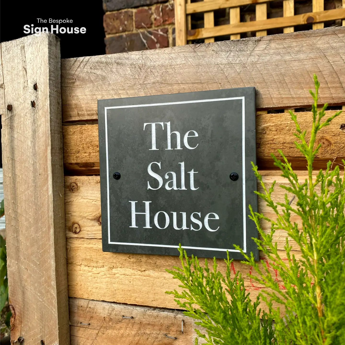 Custom slate house sign with a square border, mounted on a wooden fence, displaying the text ‘The Salt House’ in white.