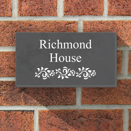 A slate house sign positioned on a wooden frame. The sign featured some floral detailing at the bottom in white. Above has the customers address engraved in white also. The sign reads Windy Ridge Lodge