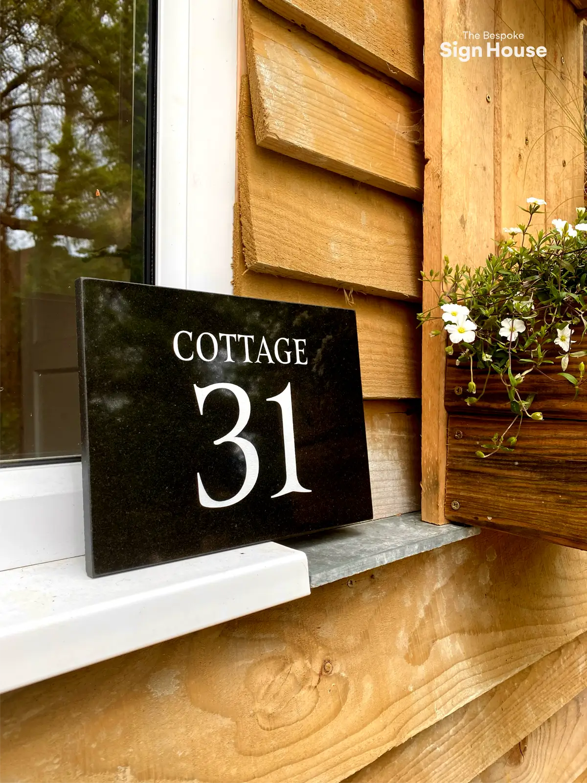 A black granite house sign with the text "COTTAGE 31" in white letters, placed on a windowsill next to a wooden wall. To the right of the sign is a wooden planter box with white flowers. In the top right corner, the text "The Bespoke Sign House" is displayed. Reflections of trees can be seen in the window glass.