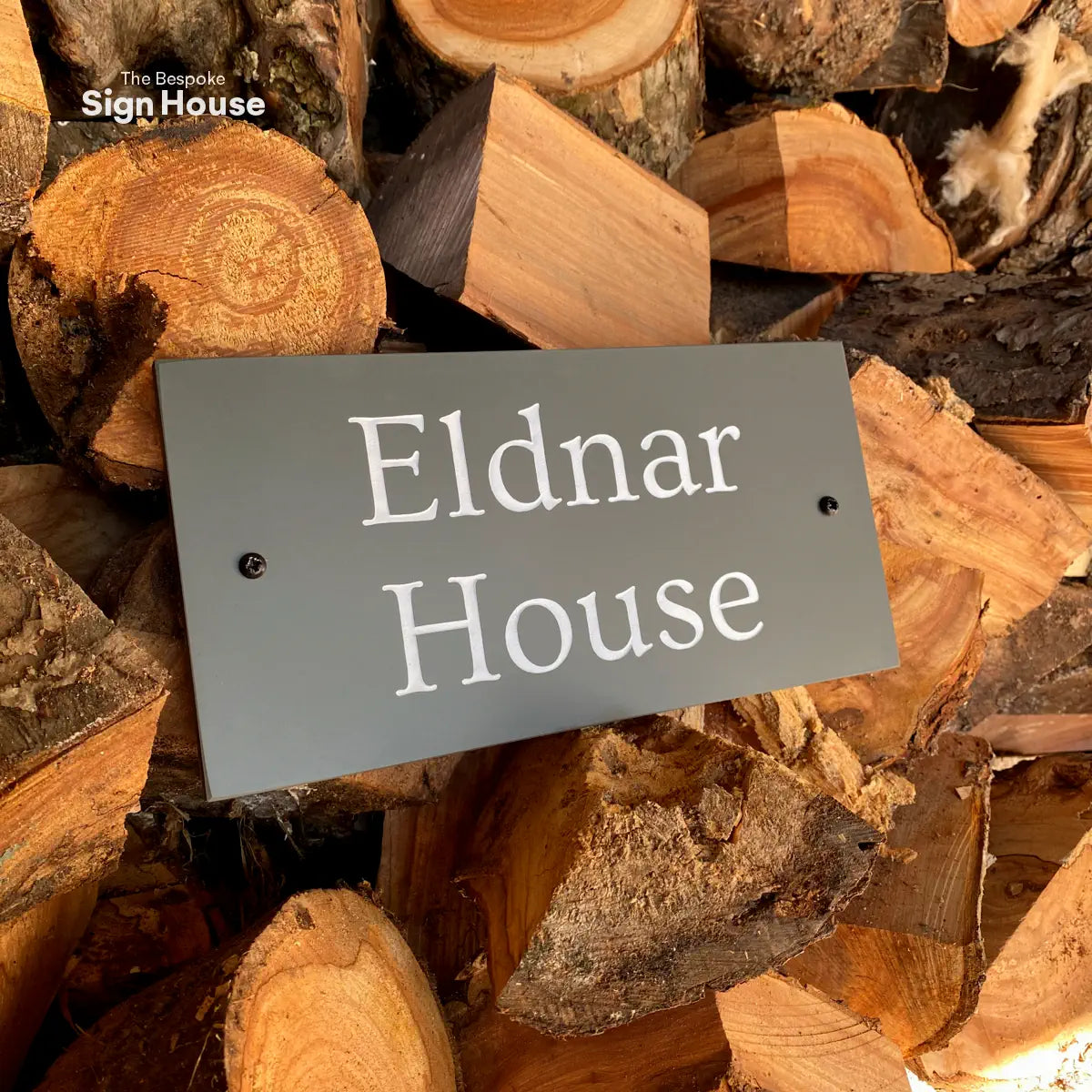 elegant slate house sign with traditional white lettering, resting on chopped up logs
