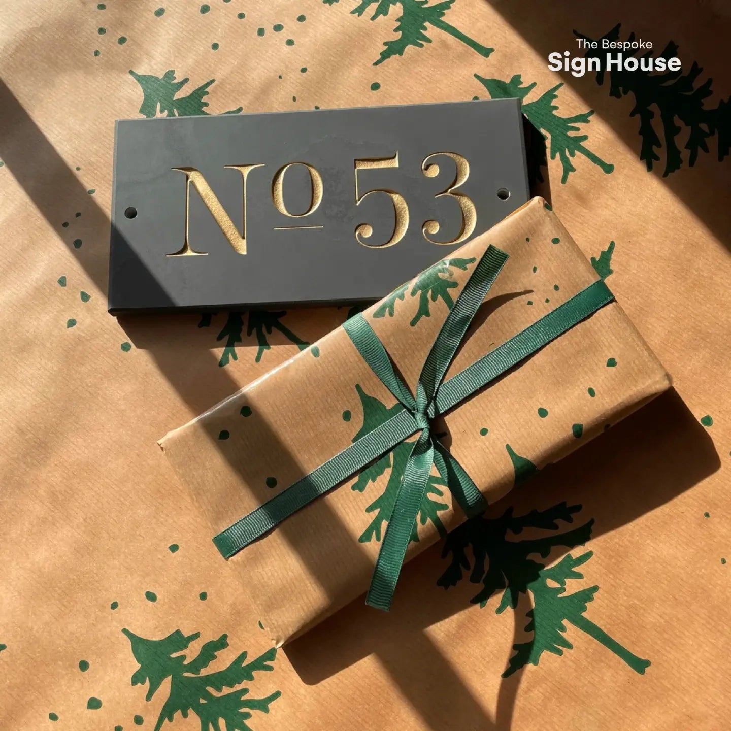 A slate house sign with the number “53” engraved in gold sits next to a neatly wrapped gift, placed on festive wrapping paper with green tree patterns.