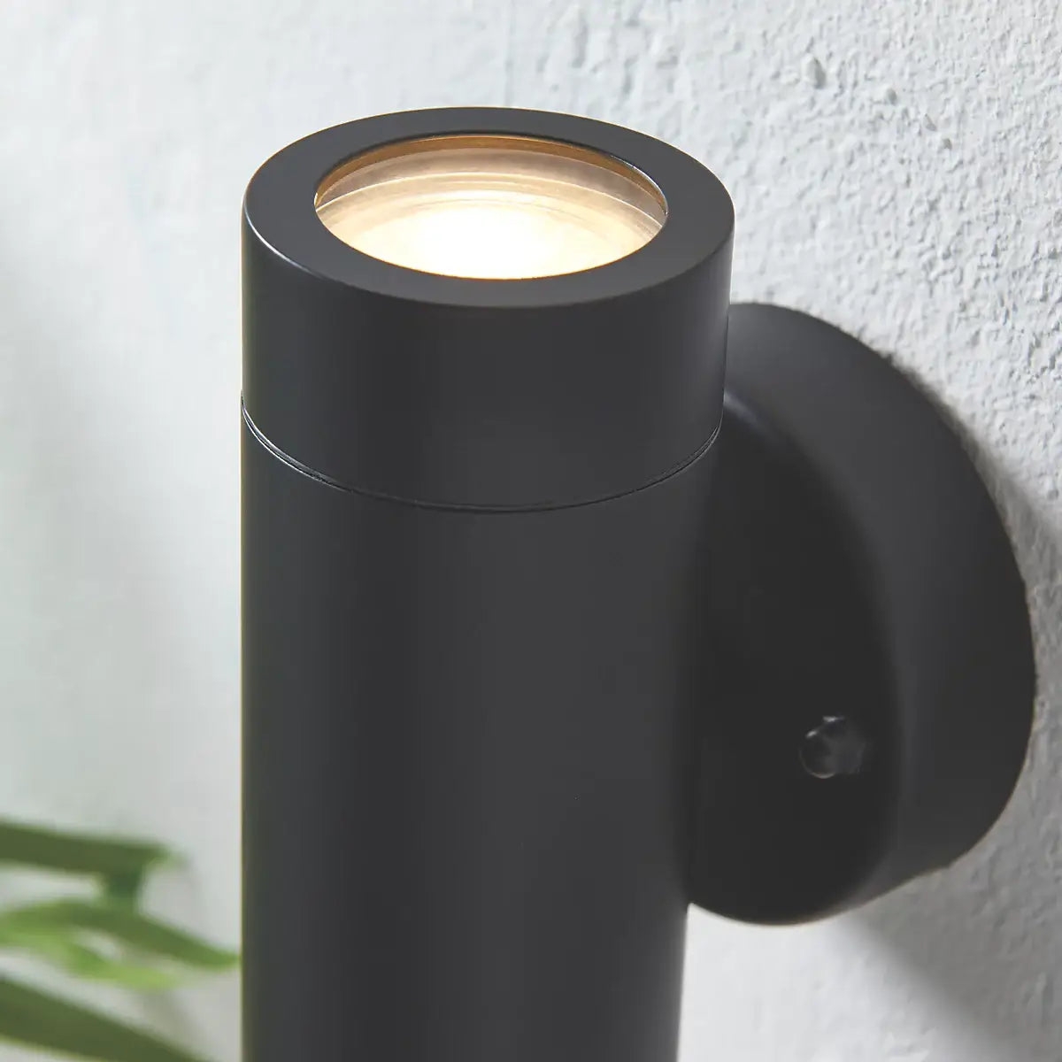 The image shows a close-up view of a sleek, cylindrical grey outdoor wall light. The focus is on the top section, with light shining upwards. The light fixture is mounted on a textured white wall, offering a modern and minimalistic appearance.