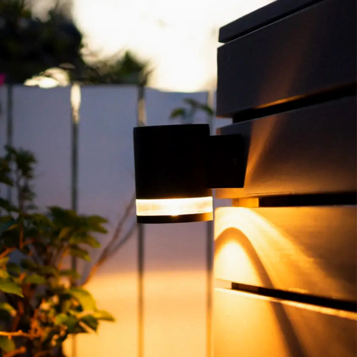 A close-up of a modern outdoor wall light, emitting a warm glow, attached to a dark wooden fence. The background features a blurred view of a white fence and some greenery. The light fixture has a sleek, cylindrical design, casting light downward and creating a cozy ambiance in the evening setting.