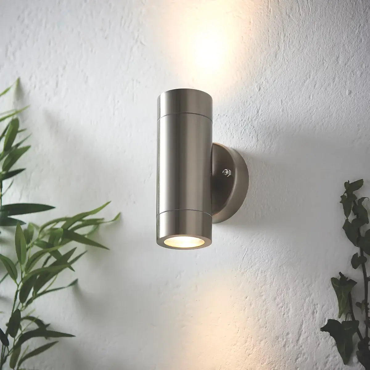 The image shows a modern silver outdoor wall light with a sleek cylindrical design. It emits both upward and downward lighting, mounted on a clean white wall with plants nearby, creating a stylish, ambient outdoor look.