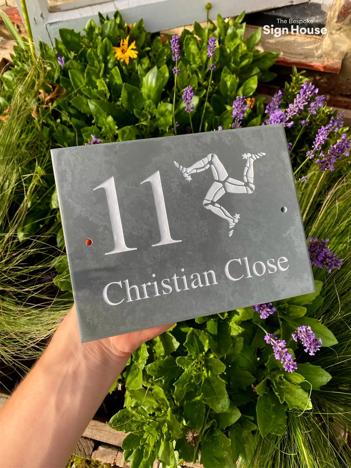 Person holding a grey slate house sign featuring the number 11, a custom name ‘Christian Close,’ and an engraved Isle of Man triskelion emblem, set against a background of lush green plants and purple flowers