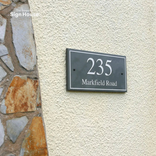 a slate house sign with a border design on a beige pebble-dashed UK house. The sign has engraved numbers and letters that are painted white