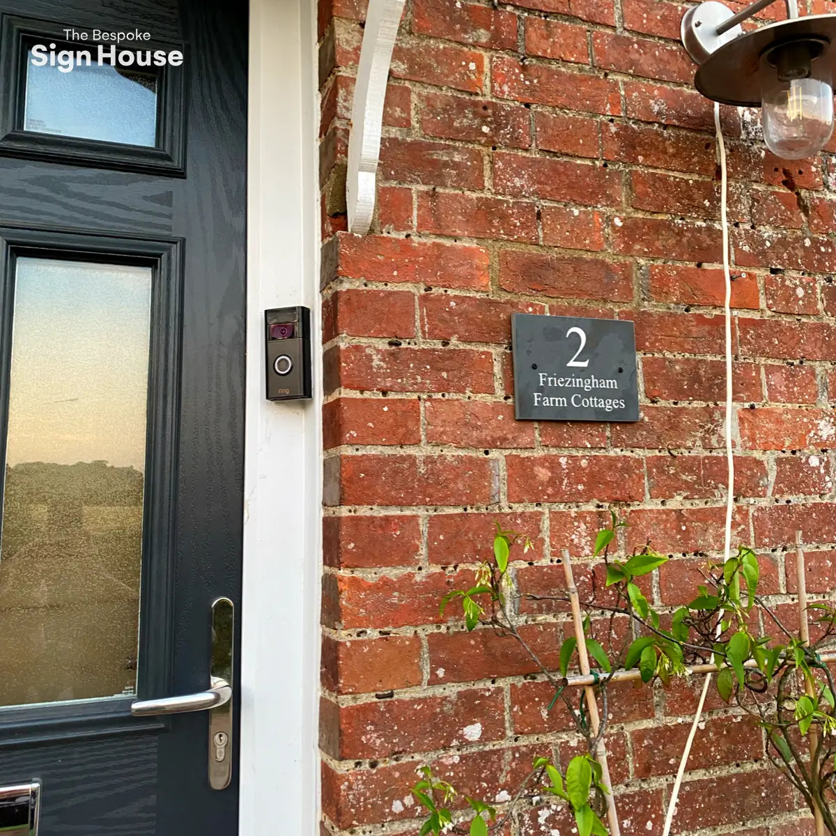 A brick wall with a black door, house sign reading "2 Friezingham Farm Cottages," and a video doorbell beside the door.