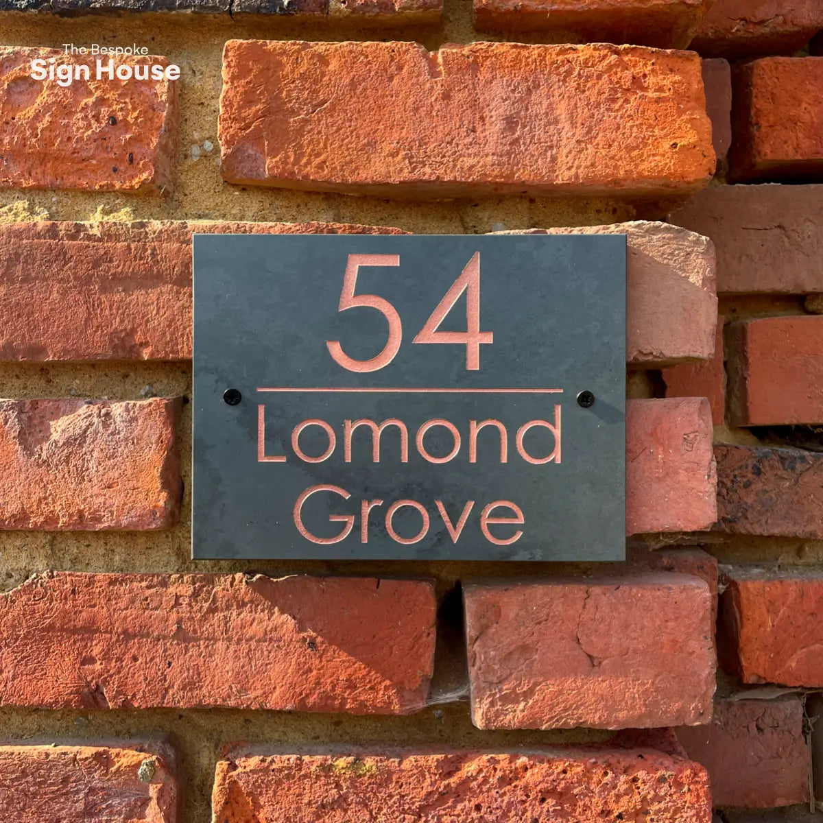 A close-up of a slate house sign reading “54 Lomond Grove” in copper lettering, mounted on a textured red brick wall.