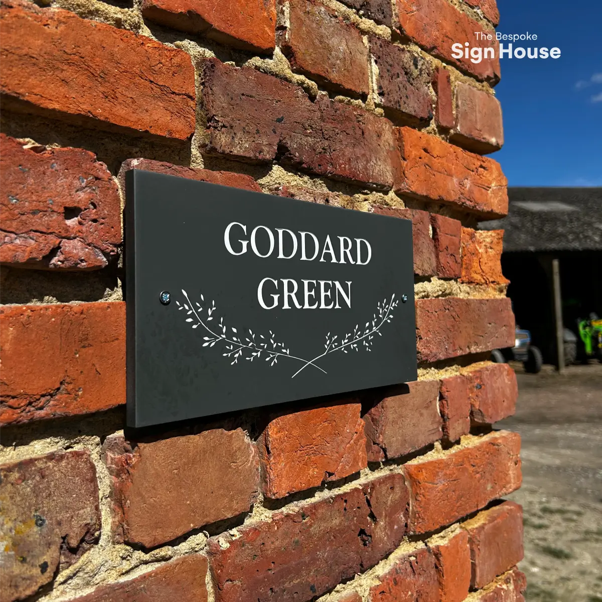 The image shows a slate house sign with the text “GODDARD GREEN” in white uppercase letters, mounted on the corner of a brick wall. Below the text is a crossed sprig design. The sign 