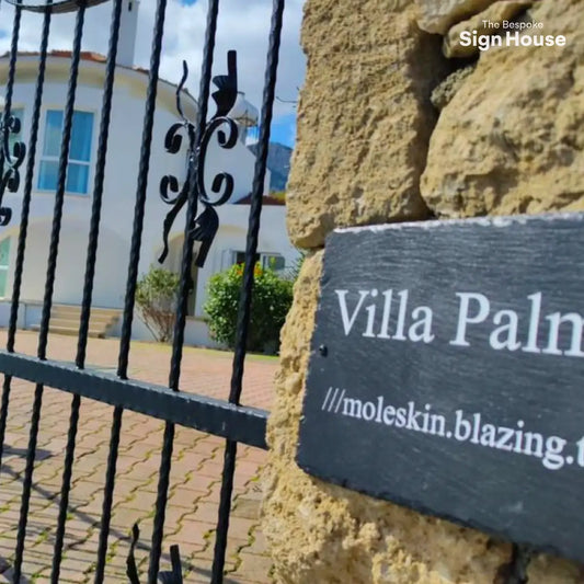 a welsh slate black sign with Villa Palma engraved onto it and painted white. The sign is on a stone wall next to black iron gates in Cyprus