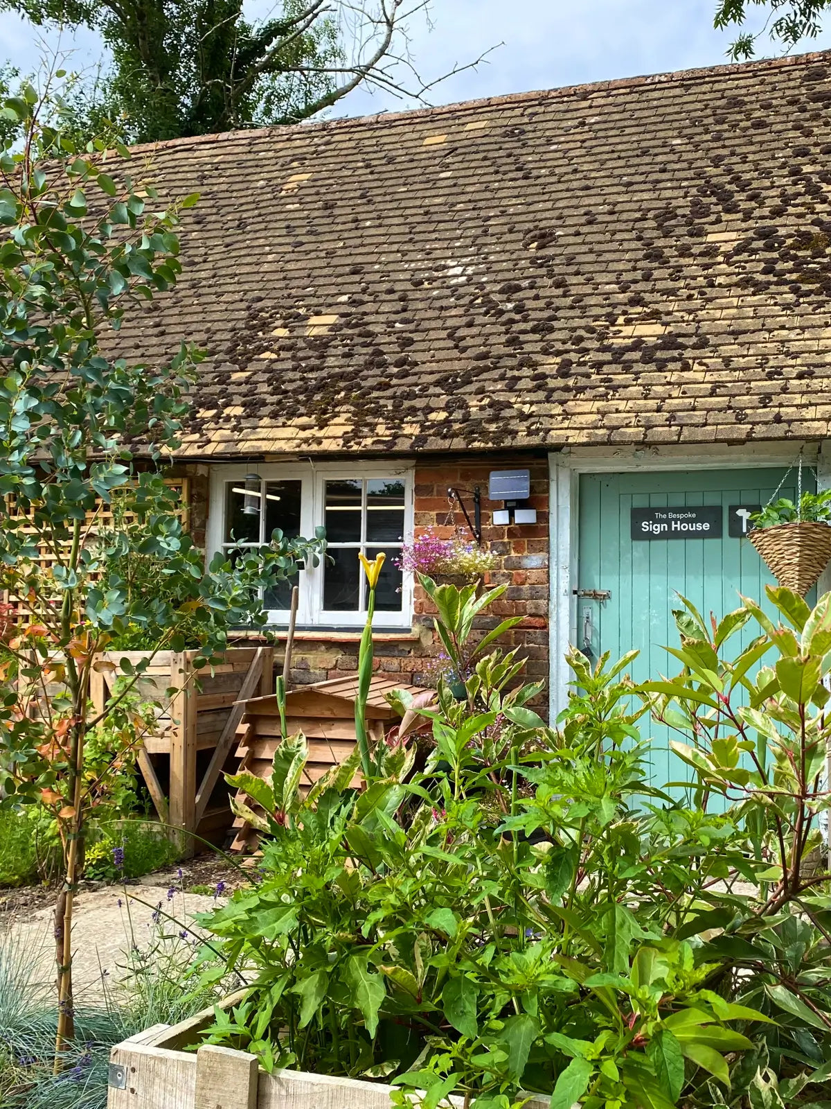 A quaint brick workshop with a sloped roof, adorned with a sign reading “The Bespoke Sign House.” The entrance has a light blue door. Surrounding the building is a lush, colorful garden with various plants and flowers, a small tree in the foreground, and wooden structures, creating a charming and vibrant atmosphere.